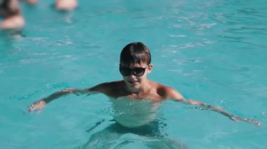 Güneş gözlüğü takan çocuk havuzda eğleniyor. Açık turkuaz su, sıcak bir yaz gününde güneşin altında parlar. Seyahat veya tatil temaları için mükemmel.