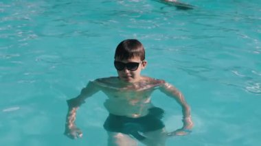 Güneş gözlüklü, mavi bilezik takan, güneşli yaz günlerinde havuzda vakit geçirmekten hoşlanan genç bir çocuk. Tatil havası, temiz su, ferahlatıcı yüzme. Yavaş çekim.