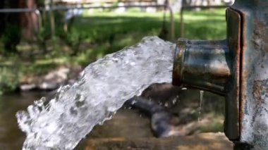 Temiz su, eski metal bir musluktan yeşil çimlerin ve ağaçların bulanık arka planına doğru akar. Bu görüntüler su kıtlığı ve korunma temalarını temsil edebilir..