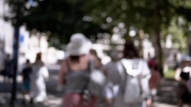 Yayalar ve ağaçlarla kaplı bir yaz caddesinin bulanık görüntüsü. Sırt çantalı iki kadın yürür, şehir hayatının anonim ve atmosferik bir temsilini oluşturur..