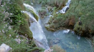 Şelaleler yosunlu yeşil bir yamaçtan aşağı akıyor ve sakin bir atmosfer yaratıyor. Turkuaz su, yemyeşil çimenler ve bitkilerle çevrili kayaların üzerinden akar.