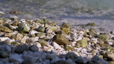 Yumuşak dalgalar bir çakıl taşı plajını yıkayıp rahatlatıcı bir ritim yaratıyor. Düz taşlar güneşin altında parıldıyor, sakin bir kıyı manzarası sunuyor..