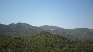 Berrak mavi gökyüzünün altındaki yeşil dağların sakin manzarası, doğanın huzurlu bir görüntüsünü sunuyor güzellik, huzur ve merak hissi uyandırıyor.