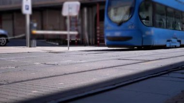 Yanımızdan geçen mavi tramvayın, tekerleklerini ve iniş takımlarını gösteren alçak açılı görüntüsü. Bulanık arka plan bir şehir sokak manzarasını ortaya çıkarıyor. Ulaşım, kentsel yaşam ve seyahat projeleri için mükemmel