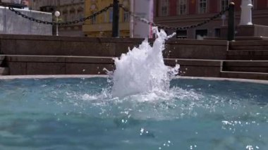 Şehir çeşmesine yakın bir yerde su sıçrıyor ve ferahlatıcı bir atmosfer yaratıyor. Arka planda bulanık binalar ve hareketler yer alıyor ve kentsel hissi artırıyor. İlgili projeler için mükemmel