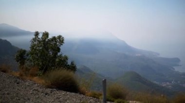 Sisli bir yaz gününde Montenegros kıyı dağlarının nefes kesici manzarası. Yeşillik yamaçları kaplıyor, dolambaçlı bir yol manzarayı kaplıyor ve Adriyatik Denizi ufka doğru uzanıyor.
