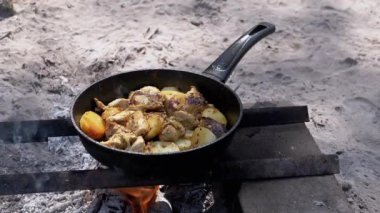 Tavuk ve patates parçaları bir kamp ateşinin üzerinde kızarıyor, lezzetli bir aroma ve kırsal alanda pişirme deneyimi yaratıyor..