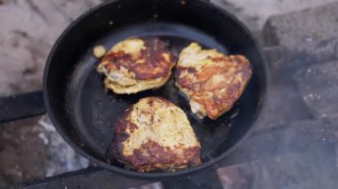 Kamp ateşi üzerinde tavada cızırdayan üç sulu tavuğun yakın çekimi. Altın-kahverengi kabuk ve yükselen duman cezbedici bir görüntü yaratır..
