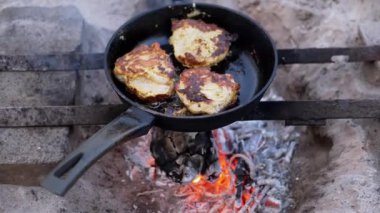 Sulu tavuk biftekleri altın kahverengiye kadar açık ateşte tavada kızartılır. Tava derme çatma bir tezgahta. Ocağın etrafında kum var. Ateş eşit derecede yanıyor. Duman yükselir.