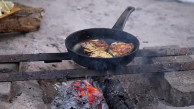 Kamp ateşinde tavada kızaran üç parça tavuk kırsal alanda yemek pişirme sahnesi yaratıyor. Tavuk pişerken duman yükselir, lezzetli bir yemek sözü verir..