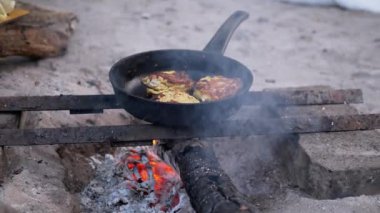 Kamp ateşinde tavada kızaran balıklı keklerin dumanlı sahnesi. İlkel bir hayatta kalma yemeği örneği..