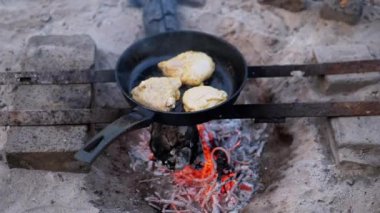 Açık ateşte tavada yağda kızartılan üç tavuk bifteği. Tava, metal çubuklardan yapılmış derme çatma bir tezgahta. Ocağın etrafında kum var. Ateş eşit derecede yanıyor, duman yükseliyor. Şey...