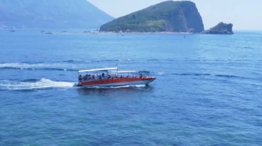 Aziz Niklas Adası yakınlarında bir turist teknesi denize açılıyor. Adanın bir plajı ve yemyeşil bitkileri var. Resimli dağlar arka planda görünür..