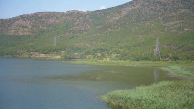 Sakin göl suyu gökyüzünü ve etrafındaki dağları yansıtır. Yeşil sazlıklar ön planda sallanıyor, arkada orman yamaçları yükseliyor. Enerji hatları arazide dolaşıyor..