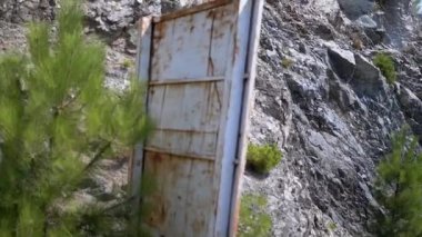 Paslı metal bir kapı kayalık bir uçurumun dibinde duruyor, doğal esnekliğin ortasında unutulmuş endüstriyi sembolize ediyor. Küçük yeşil çalılar, sert araziye tutunarak manzarayı noktalıyor. Bu çağrışımsal