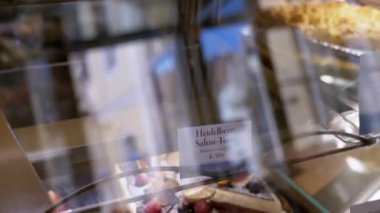 Pasta dükkânı vitrinine yakın çekimde her biri adını ve fiyatını belirten bir etiketle çeşitli pastalar ve pastalar yer alıyor. Yemek, fırın ve tatlı projeleri için ideal..