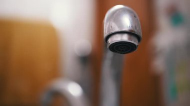 Damlayan bir musluğun yakın plan görüntüsü bulanık bir arka planda, aşağıdan görülüyor. Su damlacıkları oluşur ve düşer, su atıklarının altını çizer.