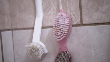 İki kullanılmış banyo fırçası fayanslı bir duvara asılmış, aşınma izleri var. Biri beyaz, diğeri sünger taşlı pembe..