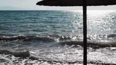 Gün batımında sıcak güneş ışığı altında deniz dalgalarını nazikçe yansıtan, bir şemsiye ile huzurlu, sakin bir sahil deniz manzarası. Ufukta dalgalar görünen fırtınalı bir deniz. Rahatla..