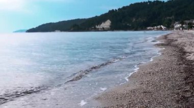 Huzurlu ve sakin bir sahil sahnesi. Yumuşak dalgalar sergiliyor. Çamurlu tepelerin altında çakıllı bir kıyı şeridine çarpıyor.