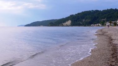 Yumuşak Dalgalar ve Güzel Bereketli Orman Arkaplanının Huzurlu Sahil Manzarası. Renkli çakıl taşları, kristal berrak su, dingin mavi gökyüzünün altındaki yemyeşil tepeler....