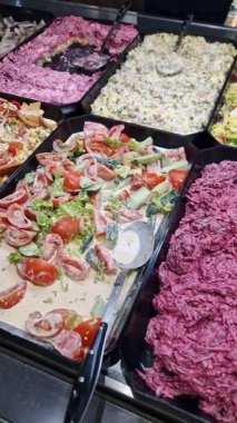 Vertical shot panning across a salad bar buffet, showing a variety of prepared dishes like beetroot salad, tomatoes with cheese, and creamy vegetable mixes in black trays at a delicatessen counter.