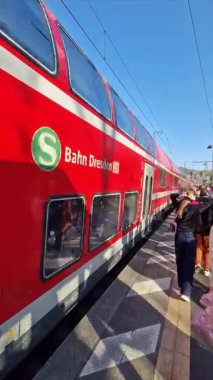 GERMANY, SAXONIAN SWITZERLAND, 2025: Vertical view of red Deutsche Bahn double-decker train departing sunny station platform, passengers wait as S1 train moves along tracks, showcasing modern German public transport and daily commutes.