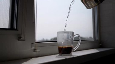 Hot water is poured from a metal kettle into a clear glass mug of coffee, sitting on a sunlit windowsill, creating a perfect morning ritual scene. Ideal for lifestyle, breakfast, and daily routine content.