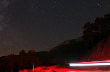 Yıldızlı gökyüzünün altından geçen kırmızı arabanın uzun süreli görüntüsü. Soyut arkaplan. Ağaçlar, ışıklar ve inanılmaz gökyüzü..