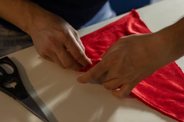 Hands of experienced worker in the handmade industry, performing cutting tasks on red fabric, on a white surface, the beginning of red hat production. Homemade sewing. Working tailor. Christmas hat.