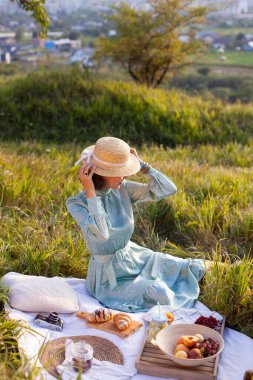 A girl in a long summer dress with short hair sitting on a white blanket with fruits and pastries and holding the straw hat. Concept of having picnic in a city park during summer holidays or weekends.