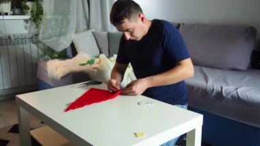 Hands of experienced worker in the handmade industry, performing cutting tasks on red fabric, on a white surface, the beginning of red hat production. Homemade sewing. Working tailor. Christmas hat.