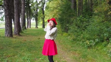 Genç kız kafasına kırmızı şapka takar ve uzun siyah saçları düzeltir. Gri kazaklı ve kırmızı etekli güzel bir kadın güneşli bir sonbahar gününde ormanda poz veriyor. Ormanda yürüyor ve dinleniyorum..