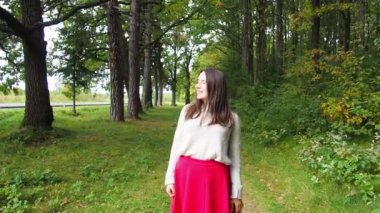 Genç kız uzun siyah saçları düzeltir. Gri kazaklı ve kırmızı etekli güzel bir kadın güneşli bir sonbahar gününde ormanda poz veriyor. Ormanda yürüyor ve dinleniyorum..