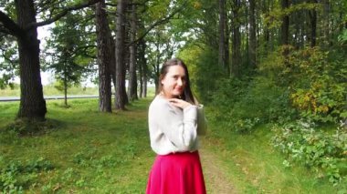Genç kız uzun siyah saçları düzeltir. Gri kazaklı ve kırmızı etekli güzel bir kadın güneşli bir sonbahar gününde ormanda poz veriyor. Ormanda yürüyor ve dinleniyorum..