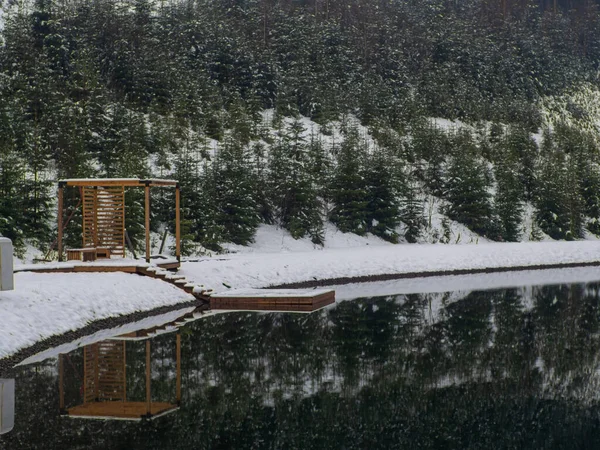 Wooden pier on the winter lake in the Carpathians, Ukraine. Landscapes with coniferous forest on a cloudy snowy day. Sights of the modern popular ski resort of Bukovel. Lake near Bukovel in winter.