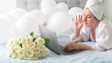 Woman in white clothes and towel on her head lying on a bed with white color balloons and bouquet of roses. Woman works at home, using her laptop, computer. Birthday, wedding. Copy space. Preparation.