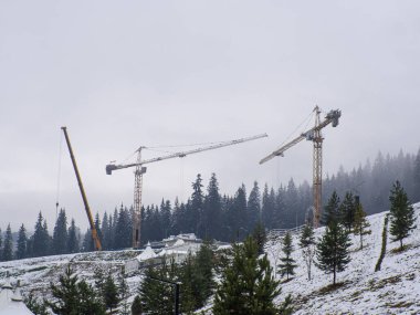 Dağlar arasında inşaat vinçleri. Kule vinçleri, gri bulutlu bir gökyüzü ve dağların arka planına karşı inşaat alanında çalışıyor. Orman ihlali şehir konsepti.