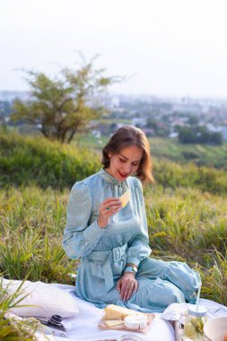 A woman in a long summer dress with short hair sitting on a white blanket with fruits and pastries and eating cheese. Concept of having picnic in a city park during summer holidays or weekends. 