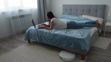 Beyaz robot süpürge, kadın yatakta yatıp dizüstü bilgisayar kullanırken yerleri temizliyor. Akıllı teknoloji konsepti. Beyaz tişörtlü ve kot pantolonlu bir kadın ev temizliğinin keyfini çıkarıyor. Model.