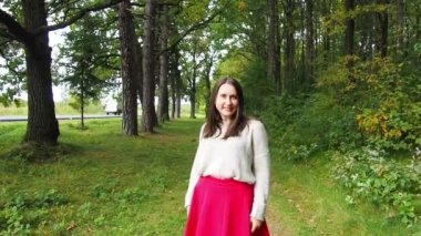 Genç kız uzun siyah saçları düzeltir. Gri kazaklı ve kırmızı etekli güzel bir kadın güneşli bir sonbahar gününde ormanda poz veriyor. Ormanda yürüyor ve dinleniyorum..