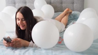 Woman in white clothes lying between white color balloons on a bed. Portrait of smiling woman using her phone with a lot of white air balloons. Happy birthday. Decoration for wedding. Copy space.