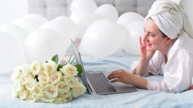 Woman in white clothes and towel on her head lying on a bed with white color balloons and bouquet of roses. Woman works at home, using her laptop, computer. Birthday, wedding. Copy space. Preparation.
