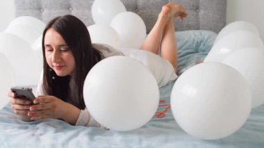 Woman in white clothes lying between white color balloons on a bed. Portrait of smiling woman using her phone with a lot of white air balloons. Happy birthday. Decoration for wedding. Copy space.