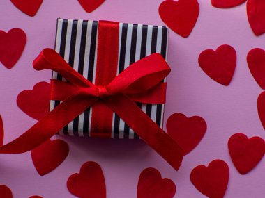 Paketlenmiş hediye paketi kırmızı kalp kağıdının arka planında, üst tarafta. Tebrik kartı, hediye. Sevgililer Günü tatili konsepti. Bir sürü kalp şeklinde kâğıdı ve yazı için yeri olan düz bir yer. 14 Şubat.