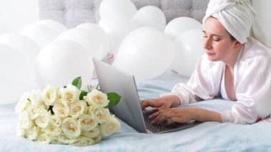 Woman in white clothes and towel on her head lying on a bed with white color balloons and bouquet of roses. Woman works at home, using her laptop, computer. Birthday, wedding. Copy space. Preparation.