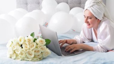 Woman in white clothes and towel on her head lying on a bed with white color balloons and bouquet of roses. Woman works at home, using her laptop, computer. Birthday, wedding. Copy space. Preparation.