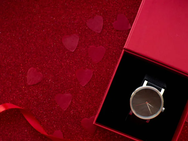 Gift box with black watch is on red heart paper background, top view. Greeting card, present. Valentines day holiday concept. Flat lay with a elegant wristwatch and space for text. February 14th.