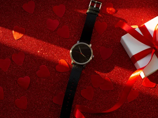 Gift box and black watch is on red heart paper background, top view. Greeting card, present. Valentines day holiday concept. Flat lay with a elegant wristwatch and space for text.