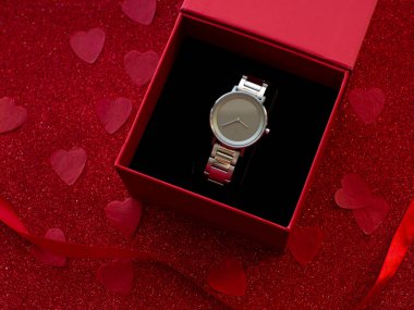 Gift box with silver watch is on red heart paper background, top view. Greeting card, present. Valentines day holiday concept. Flat lay with an elegant wristwatch and space for text. February 14th.